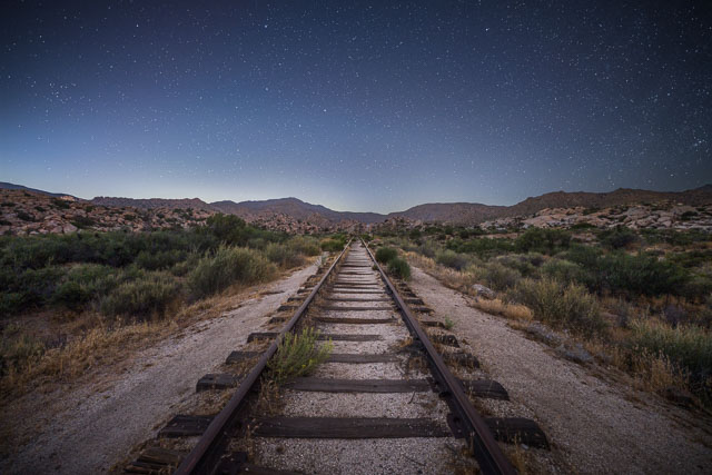 Train tracks in the center of the frame lead off into the desert and distant mountains. The night sky is full of stars.