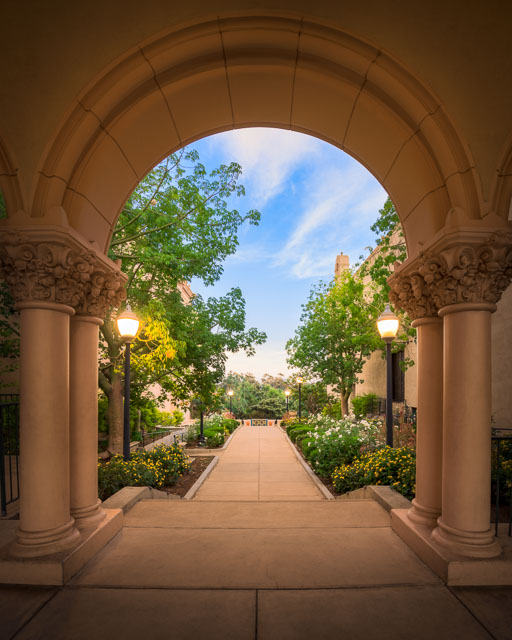 Arch surrounded by columns in Balboa Park with a pathway leading through it. Flowers line the path.