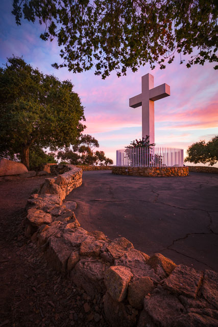 The cross in Mt Helix Park, La Mesa. It is just after sunset and there is a colorful magenta sky. A small stone wall curves into the distance leading to the cross.