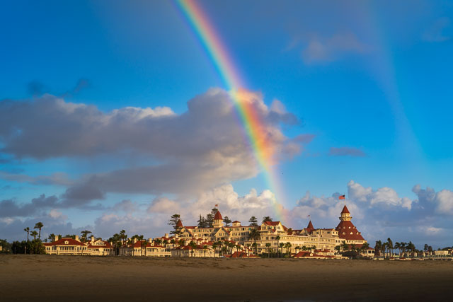 Rainbow over the Hotel Del Coronado. The arch of the rainbow comes down right in the center of the hotel. There is a blue sky and the photo was taken in the later afternoon with warm yellow light bathing the hotel.